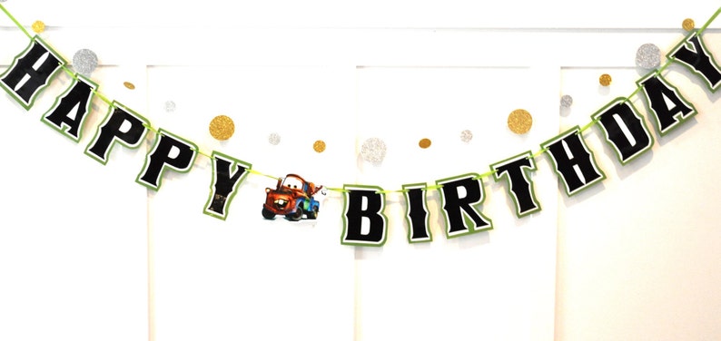 May include: A festive "HAPPY BIRTHDAY" banner with black letters outlined in white and green, strung on a green cord. The banner includes a cartoon image of a red and blue truck. Gold and silver confetti dots are scattered around the banner.
