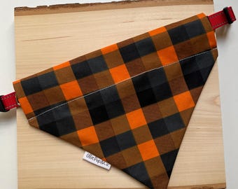 May include: A plaid dog bandana in orange, black, and brown. The bandana is triangular and has a red adjustable collar. The tag reads "OlliePop&Co.". The bandana is displayed on a wooden board.