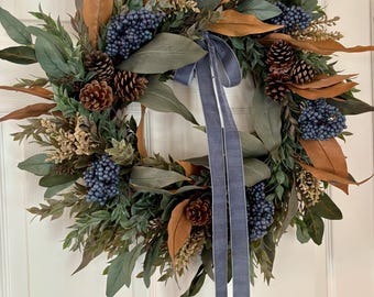 May include: A circular wreath featuring a mix of green foliage, dried brown leaves, clusters of small blue berries, and brown pinecones. A long, light blue velvet ribbon is tied at the top center, with two long tails hanging down the middle of the wreath.