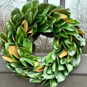 May include: A large, circular wreath composed of glossy, dark green leaves with accents of brown leaves. The wreath is displayed on a dark gray door, creating a striking contrast. The leaves are arranged in a dense, overlapping pattern, suggesting a natural, organic design.