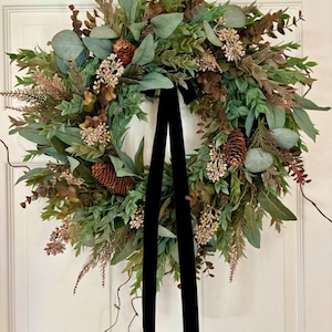 May include: A circular wreath composed of various green foliage, including eucalyptus leaves and fern-like sprigs, accented with brown pinecones and small beige floral clusters. A long, black velvet ribbon hangs vertically from the center.