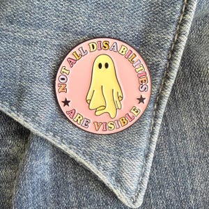 May include: A circular enamel pin with a light pink background featuring a central yellow ghost illustration. The text NOT ALL DISABILITIES ARE VISIBLE is printed in a colorful, curved font around the ghost, accented by two small black stars.