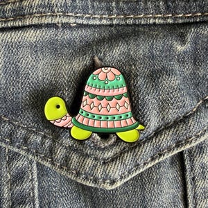 May include: A colorful turtle enamel pin with a pink and green shell, yellow head and feet, and a decorative floral design. The pin is set against a denim background.