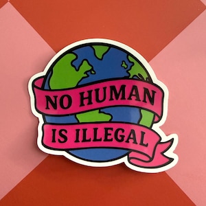 May include: A sticker featuring a stylized Earth with green continents and blue oceans. A pink banner wraps around the globe, displaying the text "NO HUMAN IS ILLEGAL" in bold white letters. The sticker is set against a red and pink background.