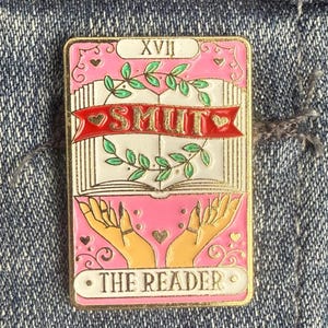 May include: A pink and gold enamel pin featuring a tarot card design. The card reads "SMUT" on a banner, with the words "THE READER" at the bottom. The pin has a gold border and is set against a denim background.