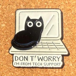 May include: A black cat-shaped pin sitting on a white laptop design. The pin has the text "DON'T WORRY I'M FROM TECH SUPPORT" in black letters. The pin is on a brown surface.