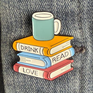 May include: A colorful enamel pin featuring a stack of books with the words "DRINK", "READ", and "LOVE". A teal mug sits atop the books. The pin is set against a denim background.