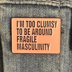 May include: A rectangular pink enamel pin with the text "I'M TOO CLUMSY TO BE AROUND FRAGILE MASCULINITY" in black lettering. The pin is set against a denim background.