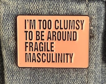 May include: A rectangular pink enamel pin with the text "I'M TOO CLUMSY TO BE AROUND FRAGILE MASCULINITY" in black lettering. The pin is set against a denim background.