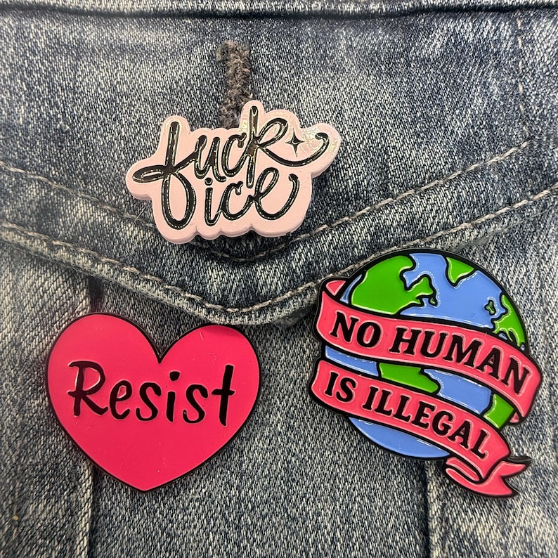 May include: Three enamel pins: a light pink pin with black cursive text reading "fuck ice", a bright pink heart-shaped pin with black text reading "Resist", and a circular pin featuring a globe with a pink banner reading "NO HUMAN IS ILLEGAL".