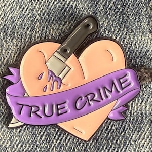 May include: A heart-shaped enamel pin with a knife piercing it. The heart is peach-colored, and the knife is black and silver. A purple banner with the words "TRUE CRIME" is wrapped around the heart. The pin is set against a denim background.