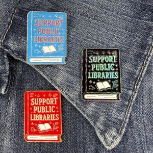 May include: Three rectangular enamel pins shaped like books. One is light blue with red text, one is black with iridescent teal text, and one is red with cream text. Each pin features the words SUPPORT PUBLIC LIBRARIES and a small open book icon.