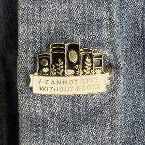 May include: A gold and black enamel pin featuring a stack of books with floral accents. The pin has a white banner with the text "I CANNOT LIVE WITHOUT BOOKS" in gold lettering. The pin is attached to a denim jacket.