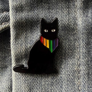May include: A black cat enamel pin featuring a rainbow-colored neck scarf. The cat is depicted in a seated position with white eyes, and the pin is attached to a blue denim fabric surface. The design is simple, featuring bold colors and lines.