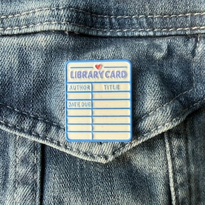 May include: A rectangular pin with a library card design. The pin features the words "LIBRARY CARD" at the top, a small red heart, and spaces for "AUTHOR", "TITLE", and "DATE DUE". The pin has a white background with blue borders.