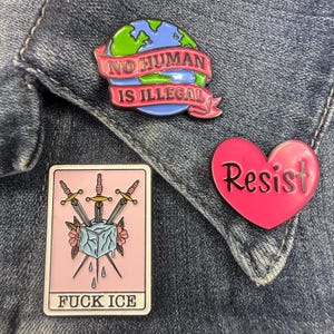 May include: Three enamel pins: a globe with a pink banner reading NO HUMAN IS ILLEGAL, a pink heart with the word Resist, and a rectangular pin featuring a tarot-style card with three swords, an ice cube, and the text FUCK ICE.