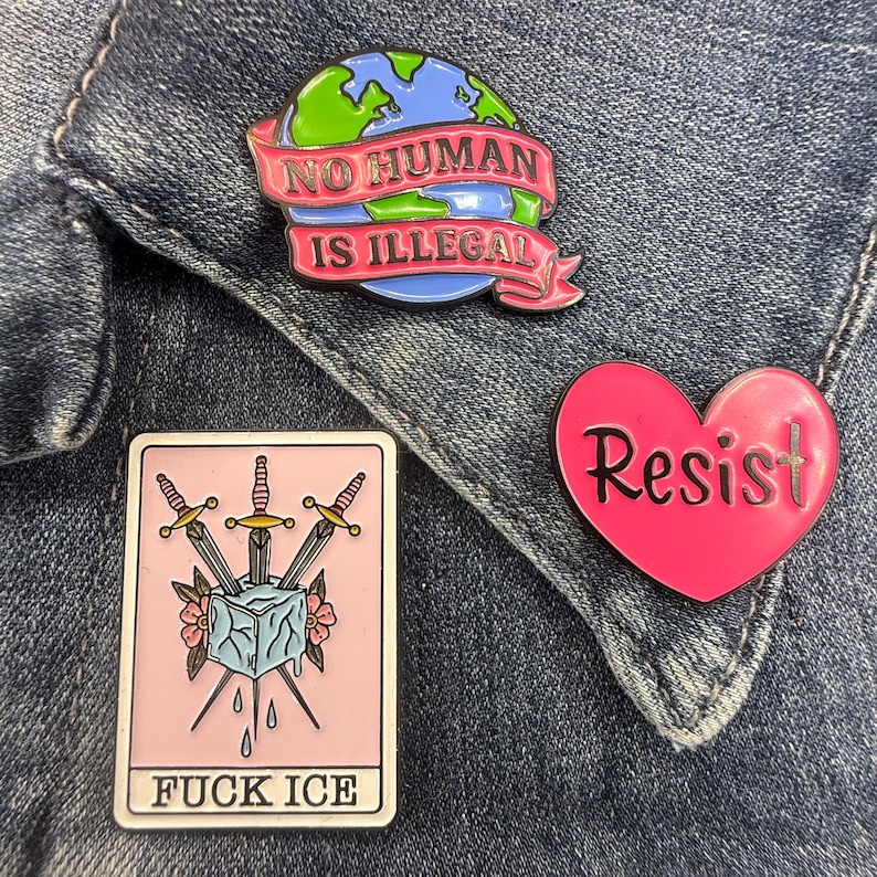 May include: Three enamel pins: a globe with a pink banner reading NO HUMAN IS ILLEGAL, a pink heart with the word Resist, and a rectangular pin featuring a tarot-style card with three swords, an ice cube, and the text FUCK ICE.