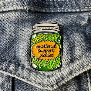 May include: A green and white enamel pin in the shape of a jar of pickles. The pin features the text "emotional support pickles" in a yellow oval. The pin is set against a blue denim background.