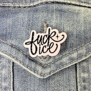 May include: A pale pink enamel pin featuring the black cursive text "fuck ice" with a small four-pointed star accent. The pin has a glossy finish and is attached to a blue denim fabric surface, showcasing a bold and clear typographic design.