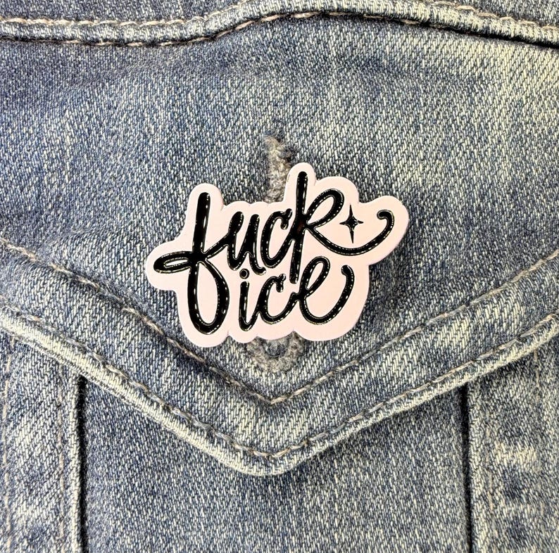 May include: A pale pink enamel pin featuring the black cursive text "fuck ice" with a small four-pointed star accent. The pin has a glossy finish and is attached to a blue denim fabric surface, showcasing a bold and clear typographic design.