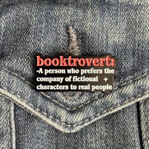 May include: A black enamel pin with a scalloped edge featuring the red text booktrovert: followed by white text that reads -A person who prefers the company of fictional characters to real people, accented with a small white four-pointed star.