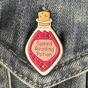May include: A colorful enamel pin in the shape of a potion bottle. The bottle is filled with pink liquid and has the words "Speed Reading Potion" on it. The pin is set against a denim background.