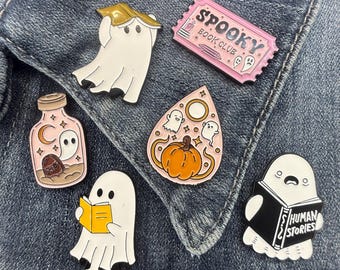 May include: A collection of six enamel pins featuring a white ghost reading a book, a pink ticket labeled Spooky Book Club, a ghost in a jar with a tombstone, a teardrop-shaped pin with a pumpkin and ghosts, and a ghost reading Human Stories.