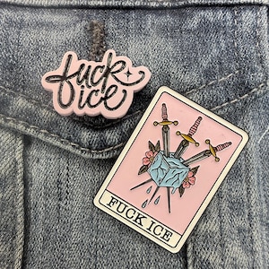 May include: Two enamel pins attached to a blue denim fabric. One pin is a light pink shape with black cursive text reading "fuck ice". The other is a rectangular tarot card design featuring a light blue ice cube pierced by three swords.