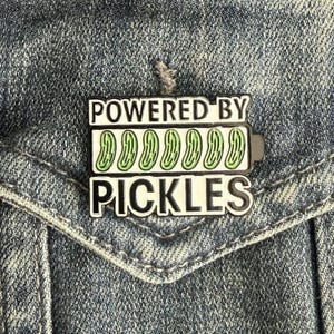 May include: A rectangular enamel pin with the text "POWERED BY PICKLES" in white letters. The pin features a battery graphic with green pickle slices. The pin is set against a denim jacket.