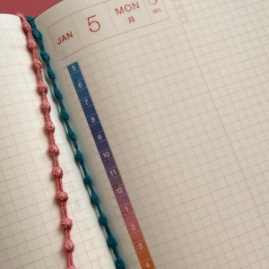 May include: Open notebook with a pink and teal beaded bookmark. The date is January 5th, Monday, with a numbered scale on the right side. The notebook pages have a grid pattern.