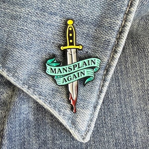 May include: A metal enamel pin featuring a vertical dagger with a yellow handle and a silver blade with red accents at the tip. A light green ribbon banner wraps around the blade, displaying the black text MANSPLAIN AGAIN in capital letters.