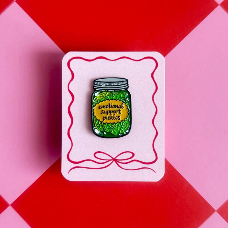May include: A pin featuring a jar of pickles with the text "emotional support pickles" on a yellow label. The pin is on a pink card with a red border and bow, set against a pink and red background.