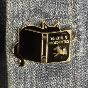 May include: Black enamel pin shaped like a cat reading a book. The book has gold trim and the title "TO KILL A MOCKINGBIRD" with a gold bird illustration. The pin is set against a denim background.