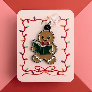 May include: A gingerbread man enamel pin reading a book, with a red bow tie. The pin is on a pink card with a red ribbon and a string of Christmas lights. The background is red and pink.