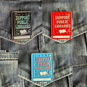 May include: Three enamel pins in the shape of books, each with the text "SUPPORT PUBLIC LIBRARIES." One pin is black with rainbow accents, one is red, and the third is light blue. Each pin features an open book illustration.