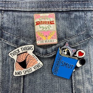 May include: Three enamel pins featuring graphic designs. One is a pink rectangular tarot card labeled SMUT THE READER. Another is a white shape with fishnet-clad legs labeled THICK THIGHS AND SMUT VIBES. The third is a blue book labeled TRASH AND ASS SMUT.