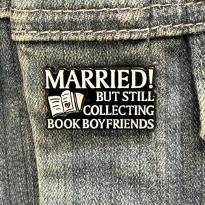May include: A rectangular enamel pin with the text "MARRIED! BUT STILL COLLECTING BOOK BOYFRIENDS" in white lettering on a black background. The pin features an open book with a heart. The pin is attached to a denim jacket.