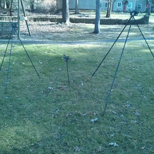 May include: Three black metal tripod stands of varying sizes on a green grassy lawn. The stands are made of sturdy metal rods and have a simple design.