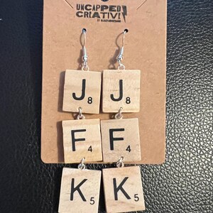 May include: A pair of handmade earrings featuring wooden Scrabble tiles spelling out the letters J, F, and K. The tiles are connected with silver-colored hooks and hang from silver-colored ear wires. The earrings are displayed on a brown card with the text "UNCAPPED CREATIVI" printed on it.