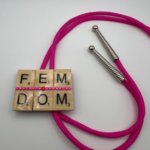 May include: A hot pink bolo tie with the word "FEMDOM" spelled out using wooden Scrabble tiles. The tiles are adorned with pink rhinestones and a single red bead. The tie features silver-colored metal tips.