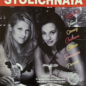 May include: Two women are shown in a black and white photo, each holding a cocktail glass. The text "Stolichnaya Flavored Russian Vodka" is in red letters at the bottom of the image. The text "Vazhna Mera (Enjoy Stoli Responsibly)" is in black letters below the Stolichnaya logo. The text "To variety. If you don't experiment, how will you know what you like?" is in black letters below the women.