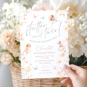 May include: A white invitation card with the text "Flutter and Twirl" and "Charlotte is turning three!" in elegant script. The card features watercolor illustrations of fairies, butterflies, and flowers in soft pink and peach tones. The invitation is held in a hand.