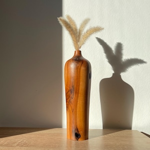 Könnte beinhalten: Eine handgefertigte Holzvase in einem warmen, rötlich-braunen Farbton. Die Vase hat eine schlanke, längliche Form mit einem abgerundeten oberen Teil und einigen kleinen, dekorativen Löchern. Sie enthält ein paar Zweige aus trockenem, federartigem Gras. Die Vase steht auf einer Holzoberfläche.