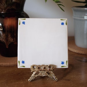 Puede incluir: Una baldosa de cerámica blanca con detalles florales azules en las esquinas, exhibida en un soporte decorativo de metal dorado. La baldosa es cuadrada y está en blanco, lista para personalizar. El soporte es ornamentado y detallado.