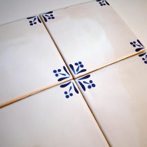 Puede incluir: Cuatro baldosas de cerámica blancas dispuestas juntas, cada una con un acabado brillante y un diseño floral azul decorativo en las esquinas y en el centro. Las baldosas son cuadradas y tienen un borde marrón claro.