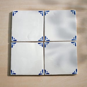 Op de afbeelding: Vier vierkante witte keramische tegels met blauwe bloemenhoekontwerpen. De tegels zijn samen gerangschikt op een licht houten oppervlak. Elke tegel heeft een glanzende afwerking en een eenvoudige, elegante esthetiek.