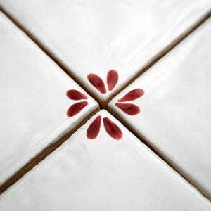 Op de afbeelding: Close-up van vier witte vierkante tegels die een kruis vormen. Elke tegel heeft een glanzende afwerking en wordt gescheiden door een dunne, bruine voeglijn. Een bordeauxrood bloemmotief bevindt zich in het midden.