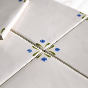 Könnte beinhalten: Nahaufnahme von weißen Keramikfliesen mit glänzender Oberfläche. Jede Fliese zeigt ein handgemaltes Blumendesign mit blauen Blumen und grünen Blättern. Die Fliesen sind zusammen angeordnet.