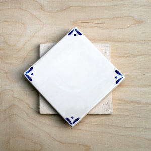 May include: A white ceramic tile with a diamond shape, featuring blue corner accents, rests atop a smaller, square, beige tile. The tiles are set against a light wood grain background, creating a contrast in textures and colours.