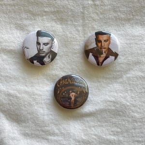 May include: Three round button pins with different images. The first pin features a black and white photo of a man with short dark hair. The second pin features a black and white photo of a man with short dark hair wearing a white shirt. The third pin features a black and white photo of a woman with long dark hair wearing a white dress. The text "Cosa Nostra" is printed in gold on the third pin.